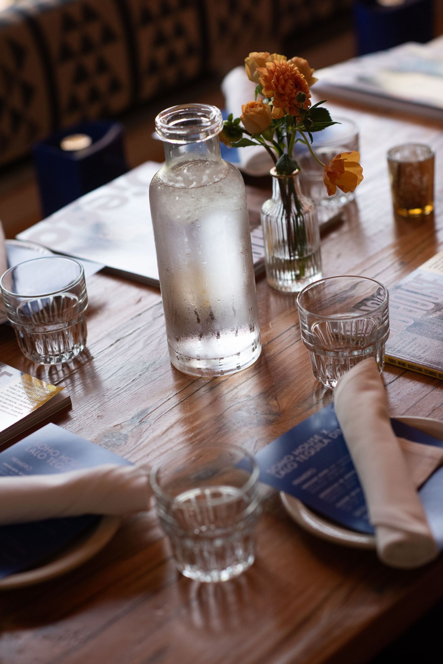 A rustic restaurant table set with glass water bottles, small drinking glasses, and orange flowers in a vase on wooden surface.