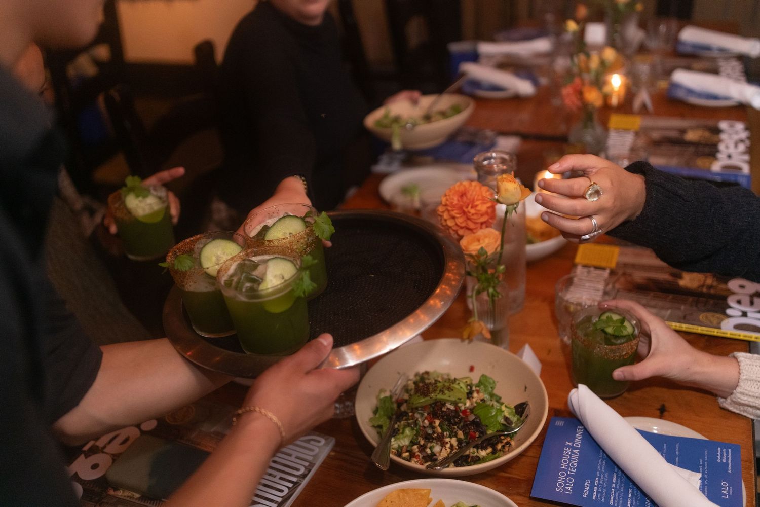 Hands reach for dishes and drinks at a dimly lit dinner table with green drinks and fresh salads.
