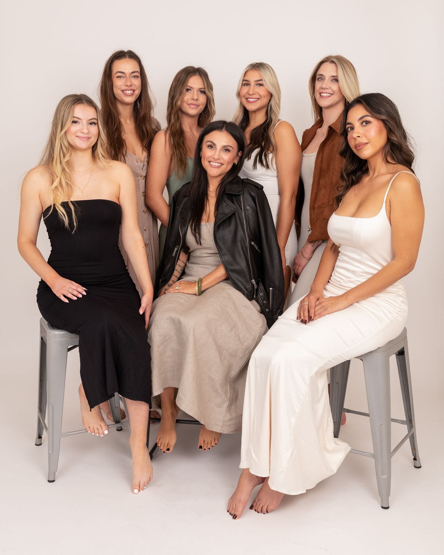 Group of seven women in casual attire posing together on metal stools in a minimal studio setting.