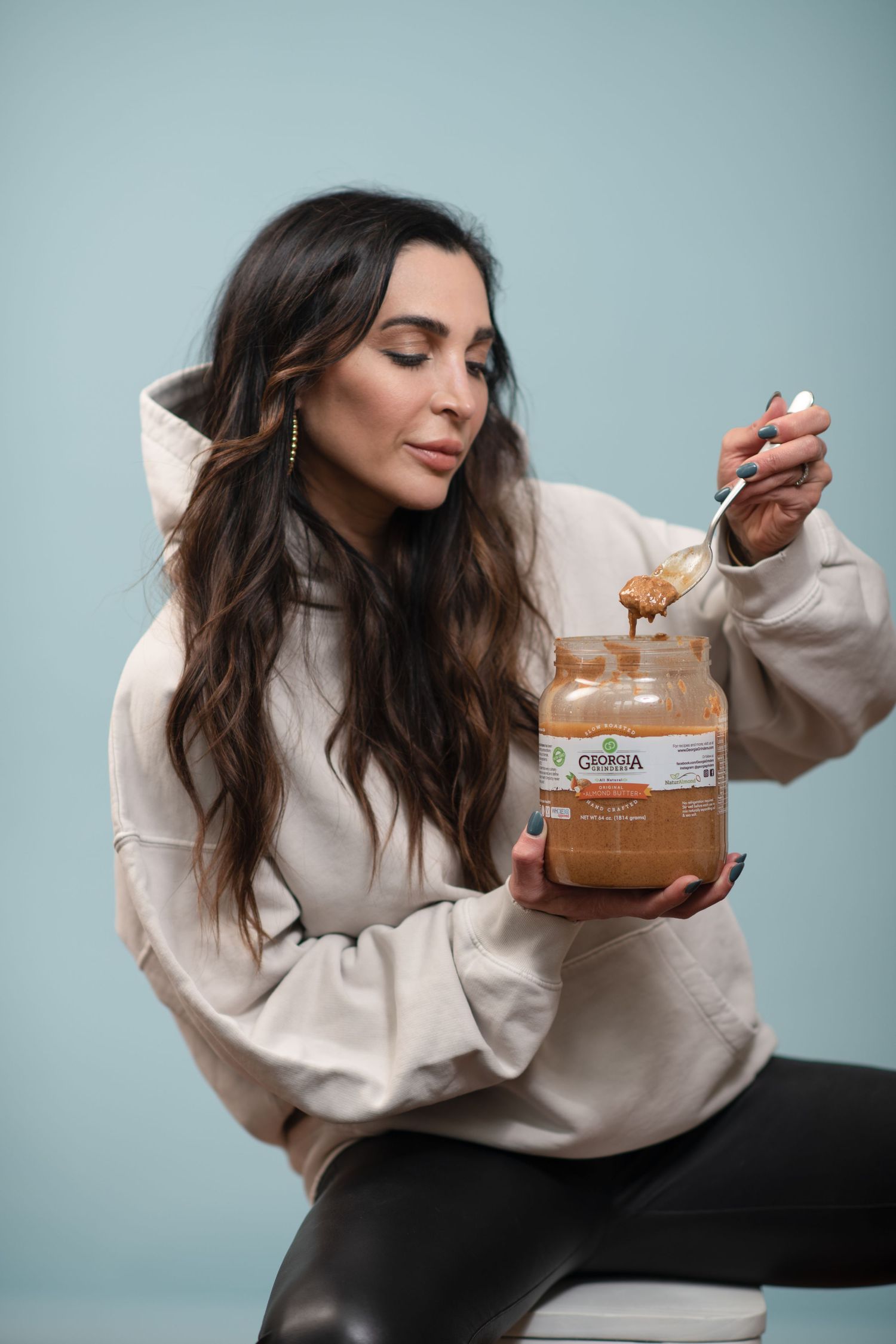 Woman in beige sweatshirt holding large jar of creamy spread while posing against light blue background.