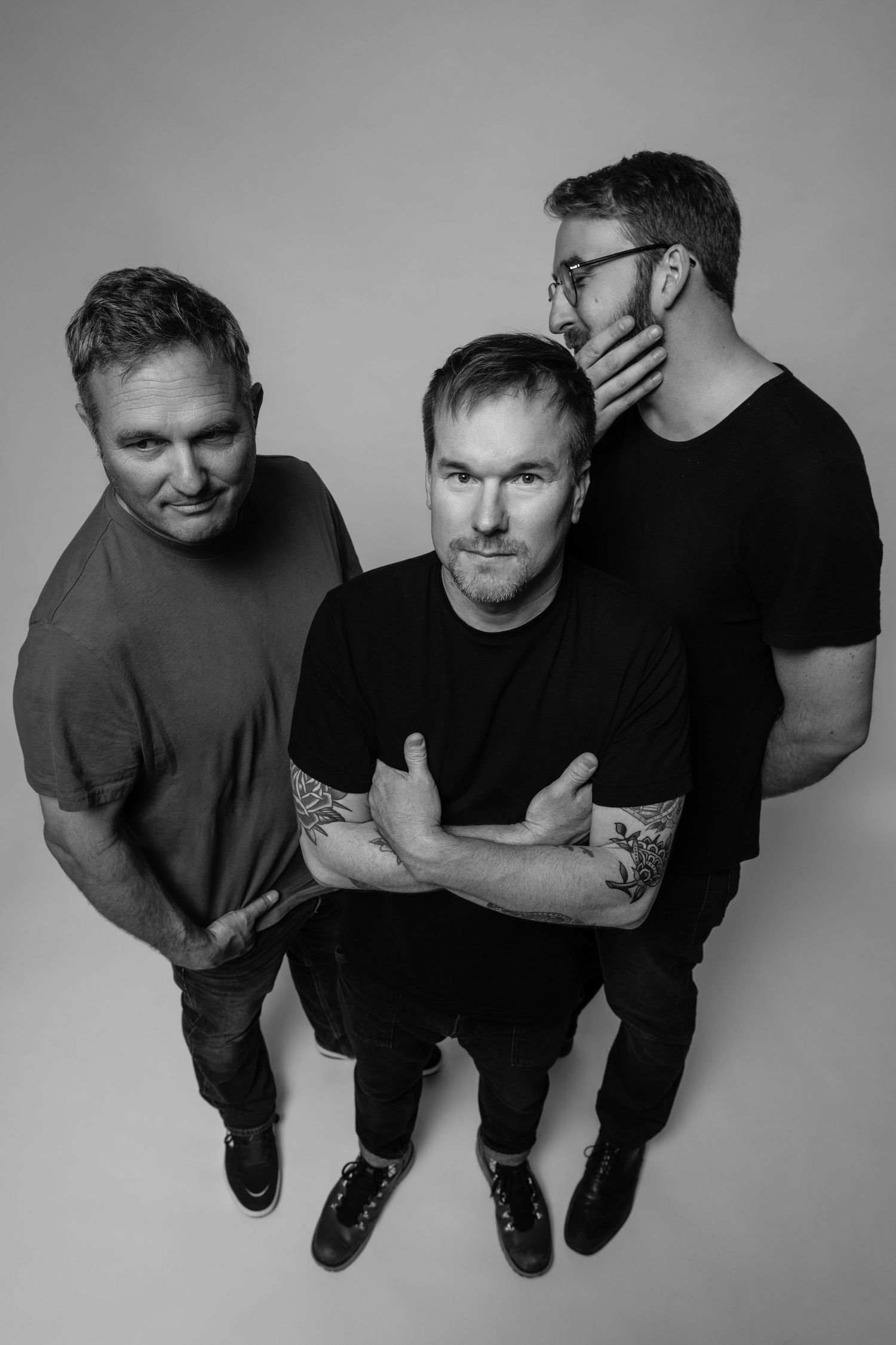 Black and white portrait of three men standing together in a minimalist studio setting against white background.
