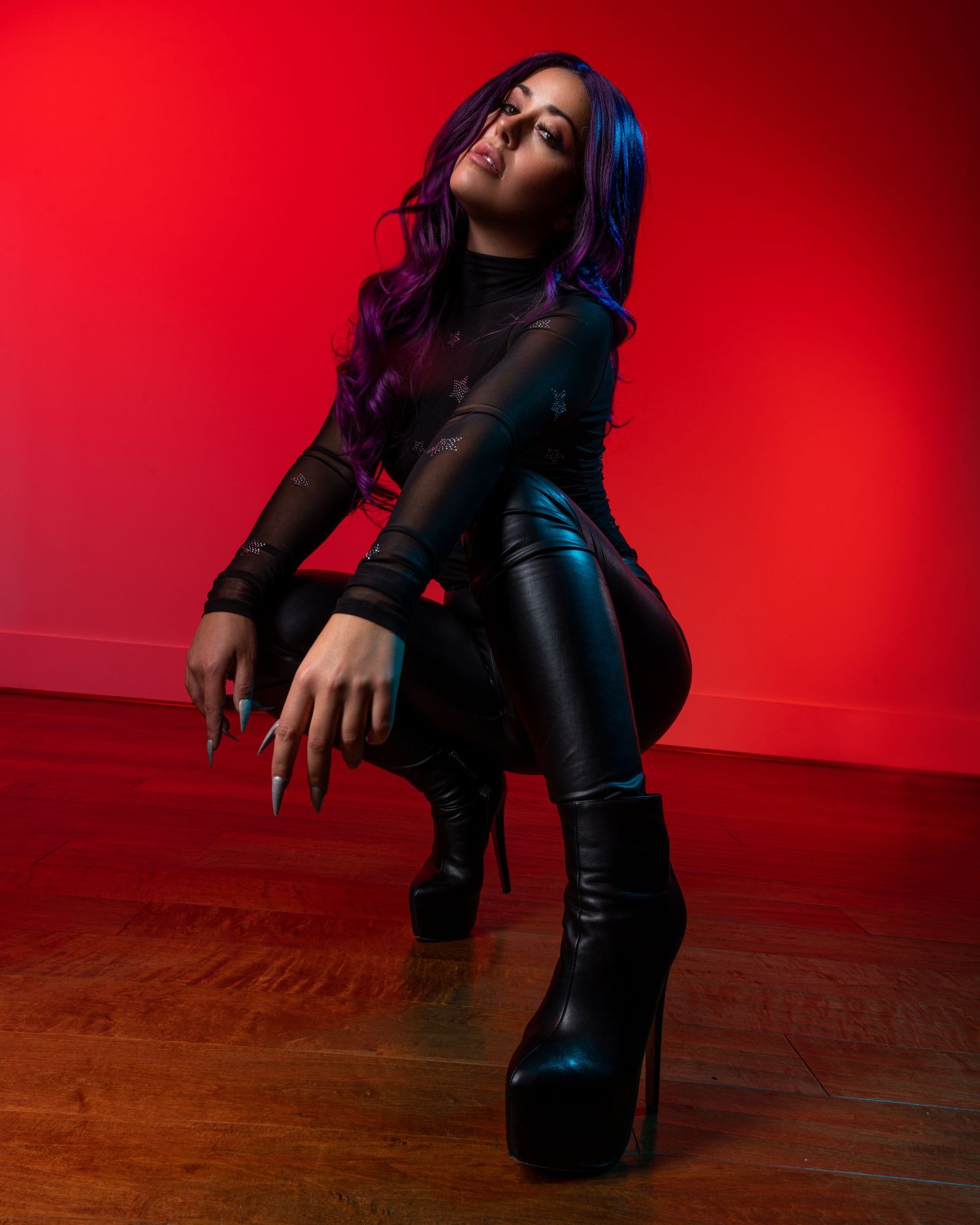 Figure in black mesh and leather outfit with purple hair poses dramatically on floor against vibrant red backdrop.