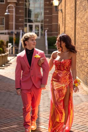 A couple in vibrant formal attire walks together on a brick sidewalk, wearing a pink suit and sparkly red dress.