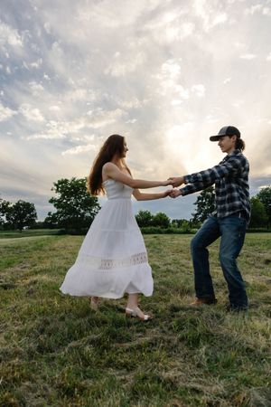 A couple dances in a green field at sunset, woman in white dress, man in plaid shirt and cap holding hands.