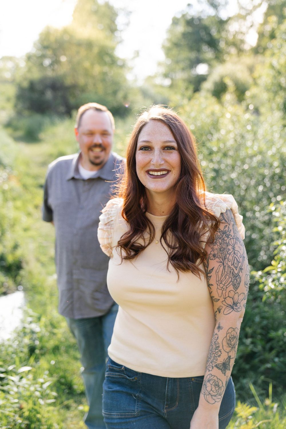 Smiling woman with tattoos and long brown hair poses outdoors with a man in a lush green setting.