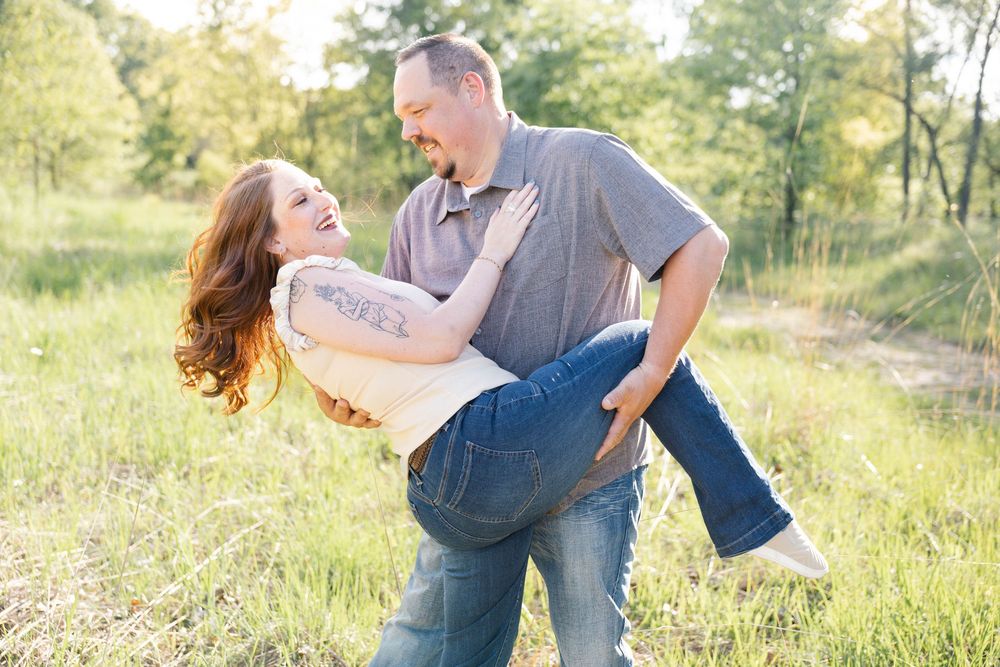 Man carrying laughing woman in his arms outdoors in a green field during a couples engagement photo session.