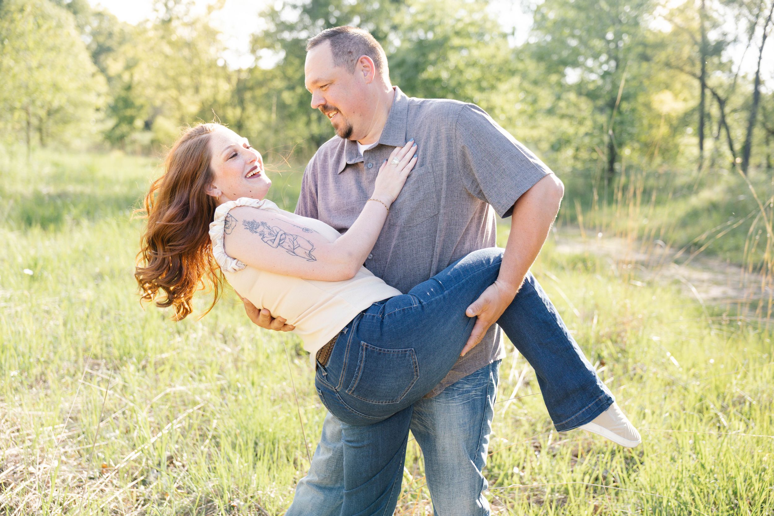 Man carrying laughing woman in his arms outdoors in a green field during a couples engagement photo session.
