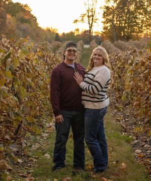 A couple stands together in a vineyard at sunset during autumn with golden light filtering through the trees.