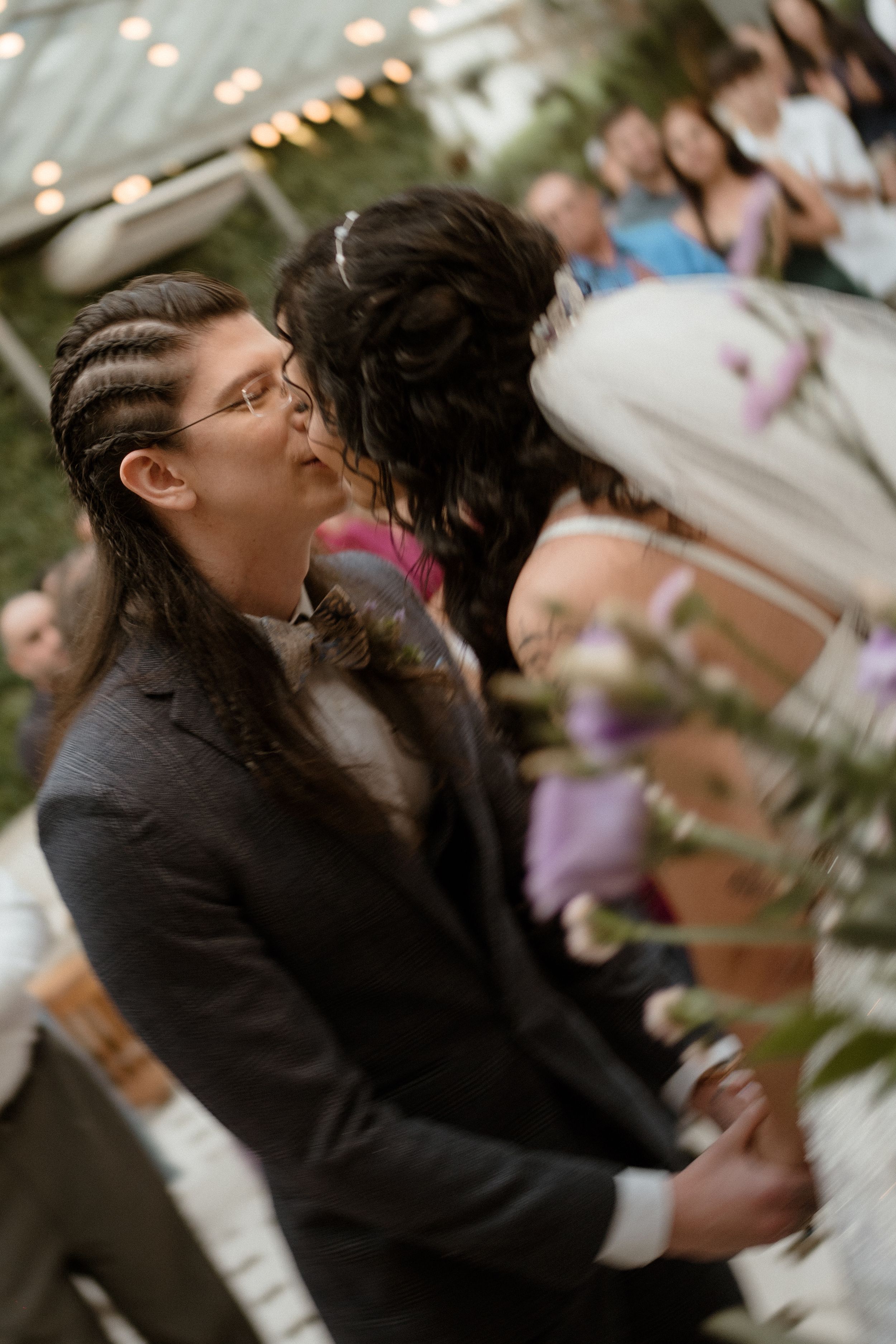 A romantic first kiss during an outdoor wedding ceremony with guests looking on.