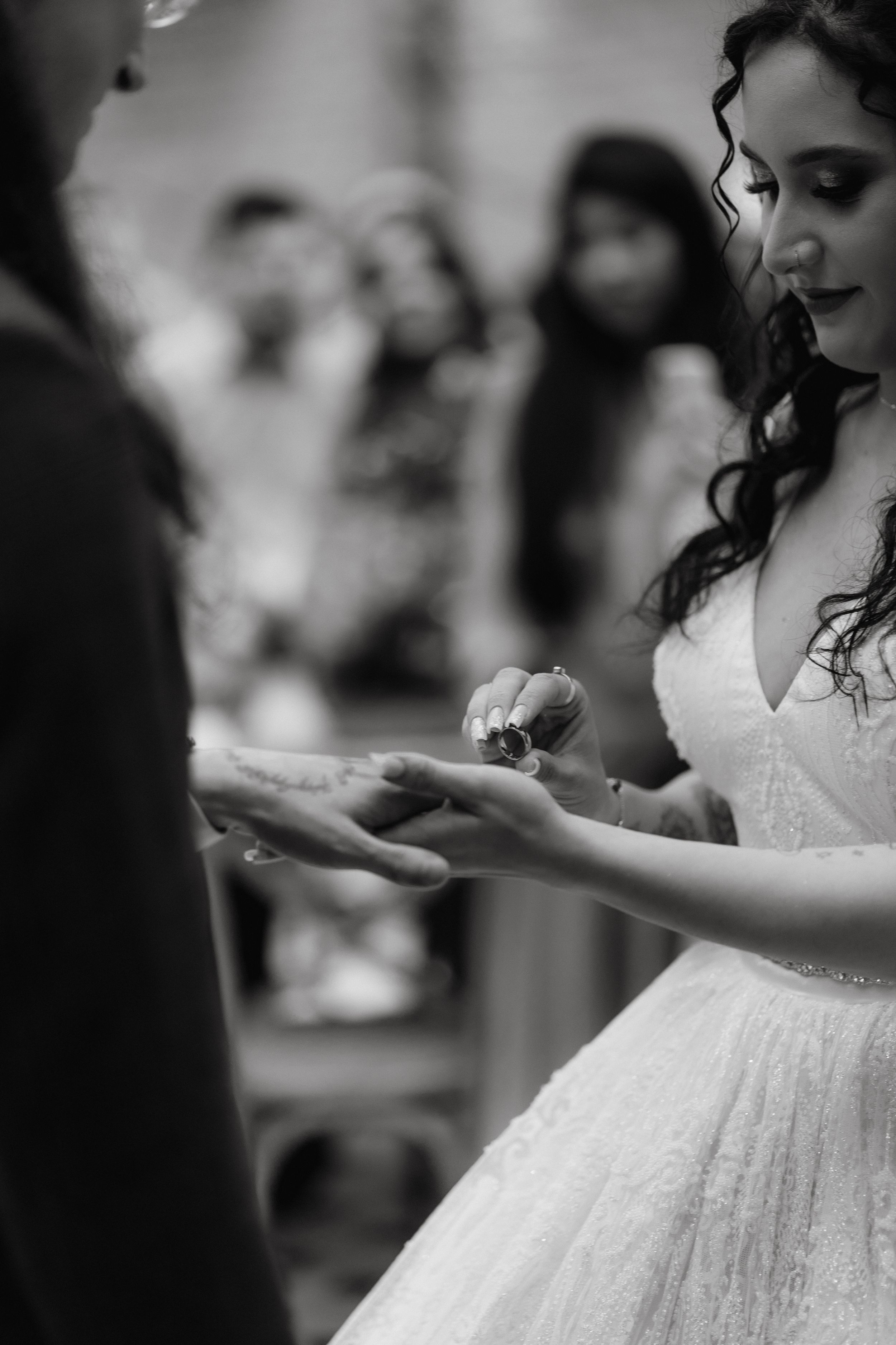 Black and white photo of a bride in a lace dress exchanging rings during wedding ceremony.