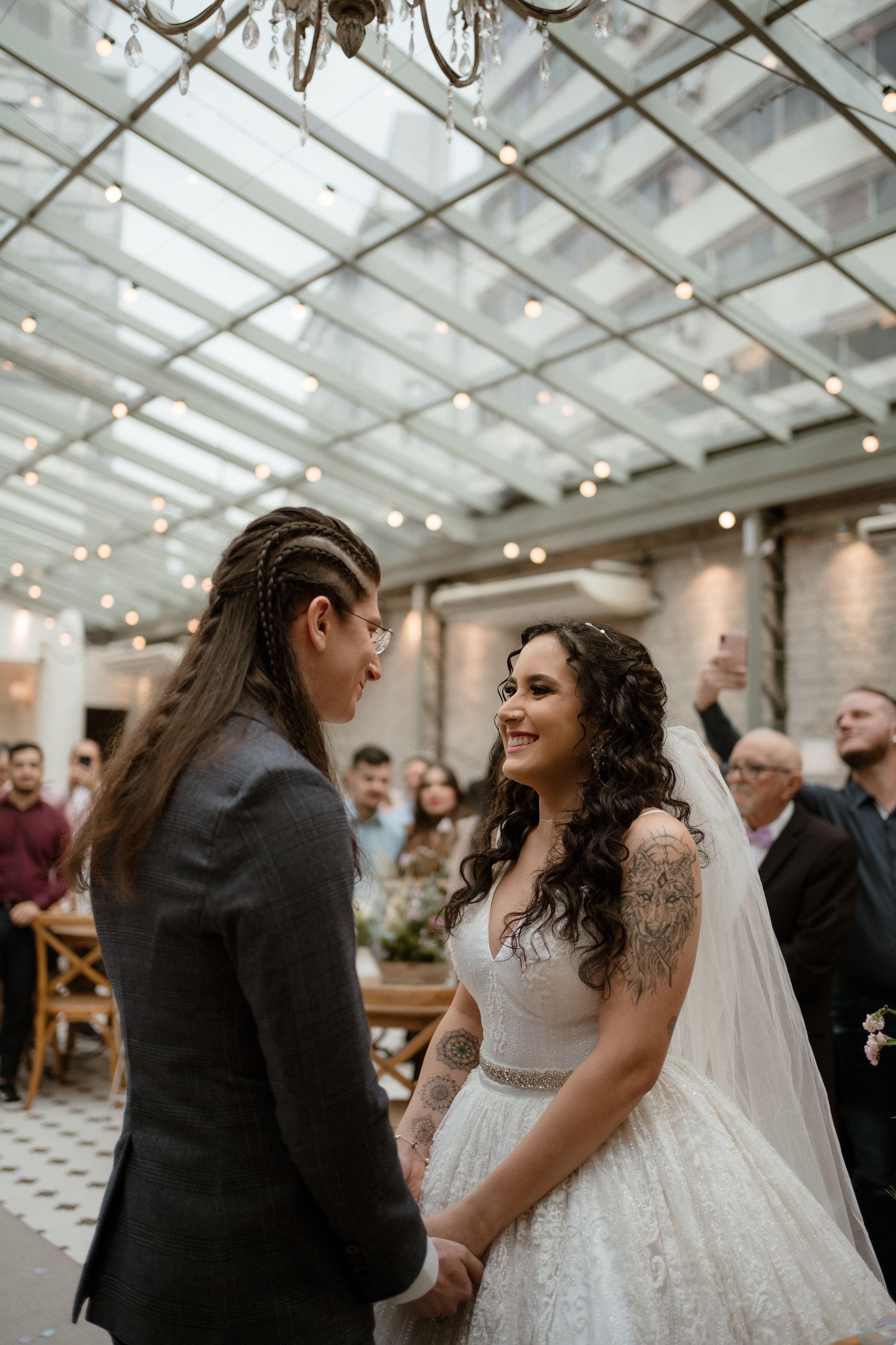 Couple shares tender moment under glass ceiling and string lights during indoor wedding ceremony.