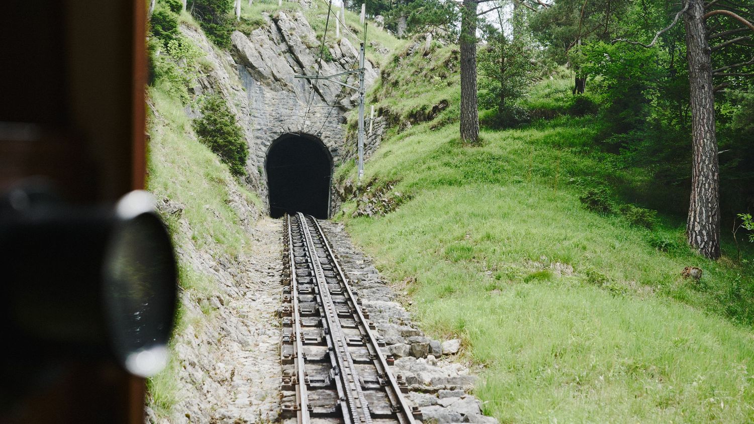 Train tracks leading into a dark tunnel surrounded by lush green hillside and pine trees.