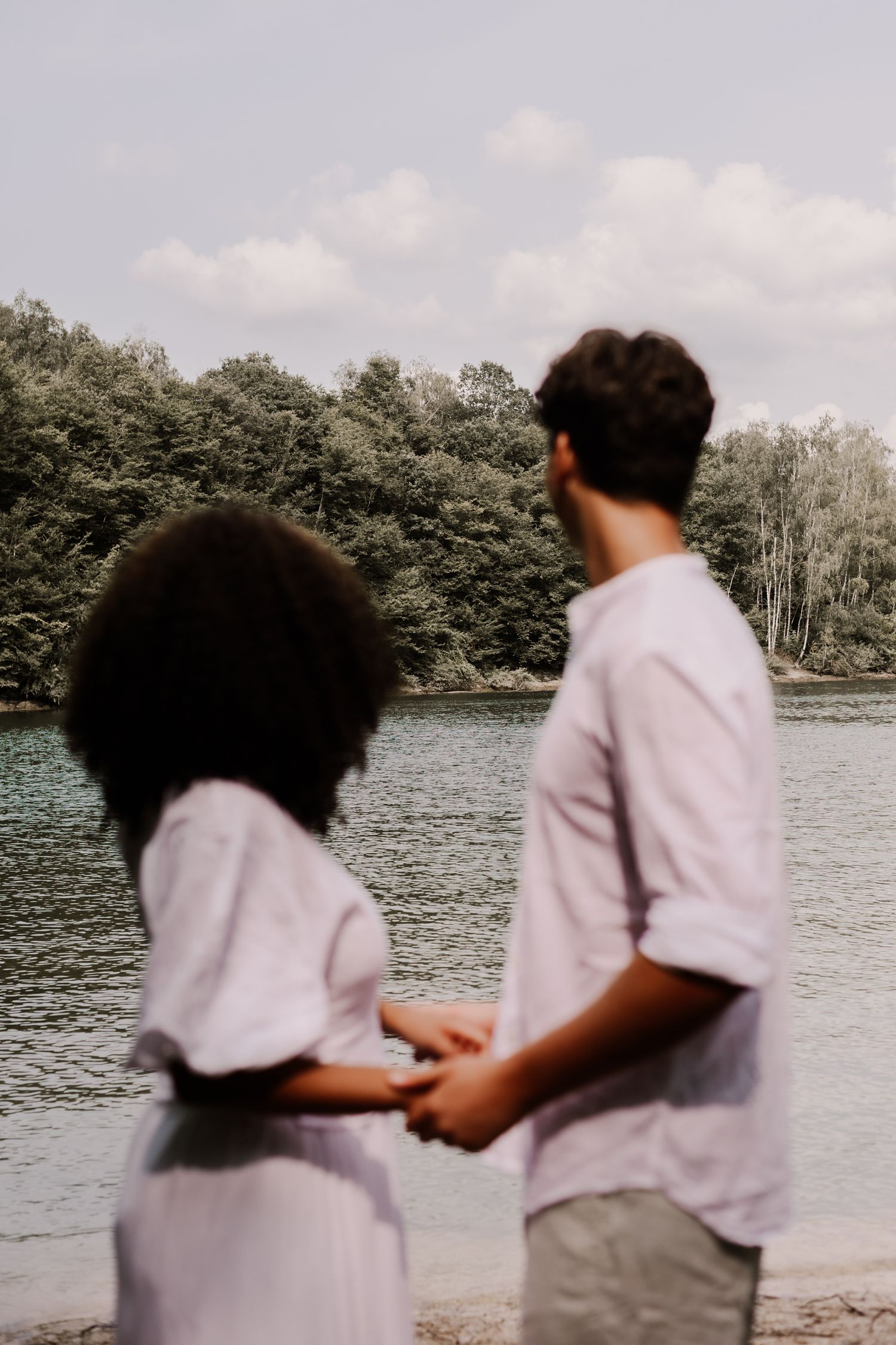 Two people in light colored shirts stand together looking out at a tranquil lake surrounded by trees on a cloudy day.