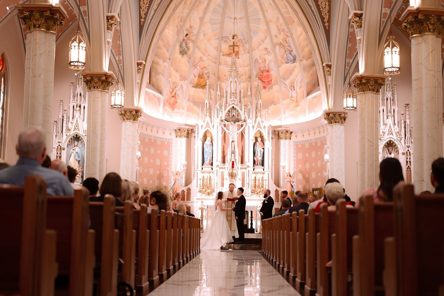 Elegant wedding ceremony inside ornate Catholic church with gilded altar, frescoed ceiling, and guests seated in wooden pews.