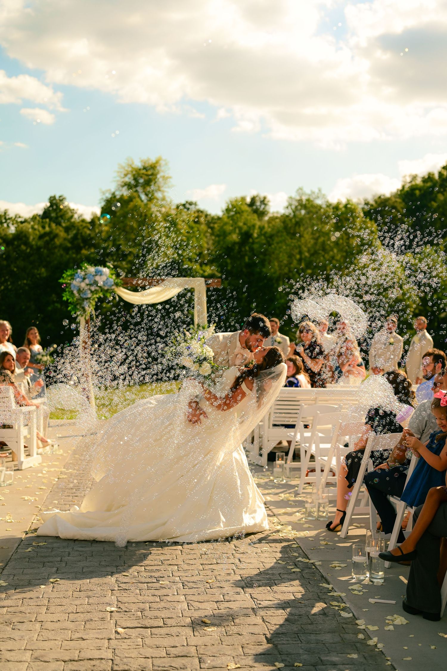 Bride and groom walk between lush green and burgundy floral arrangements on white pedestals in elegant wedding ceremony.