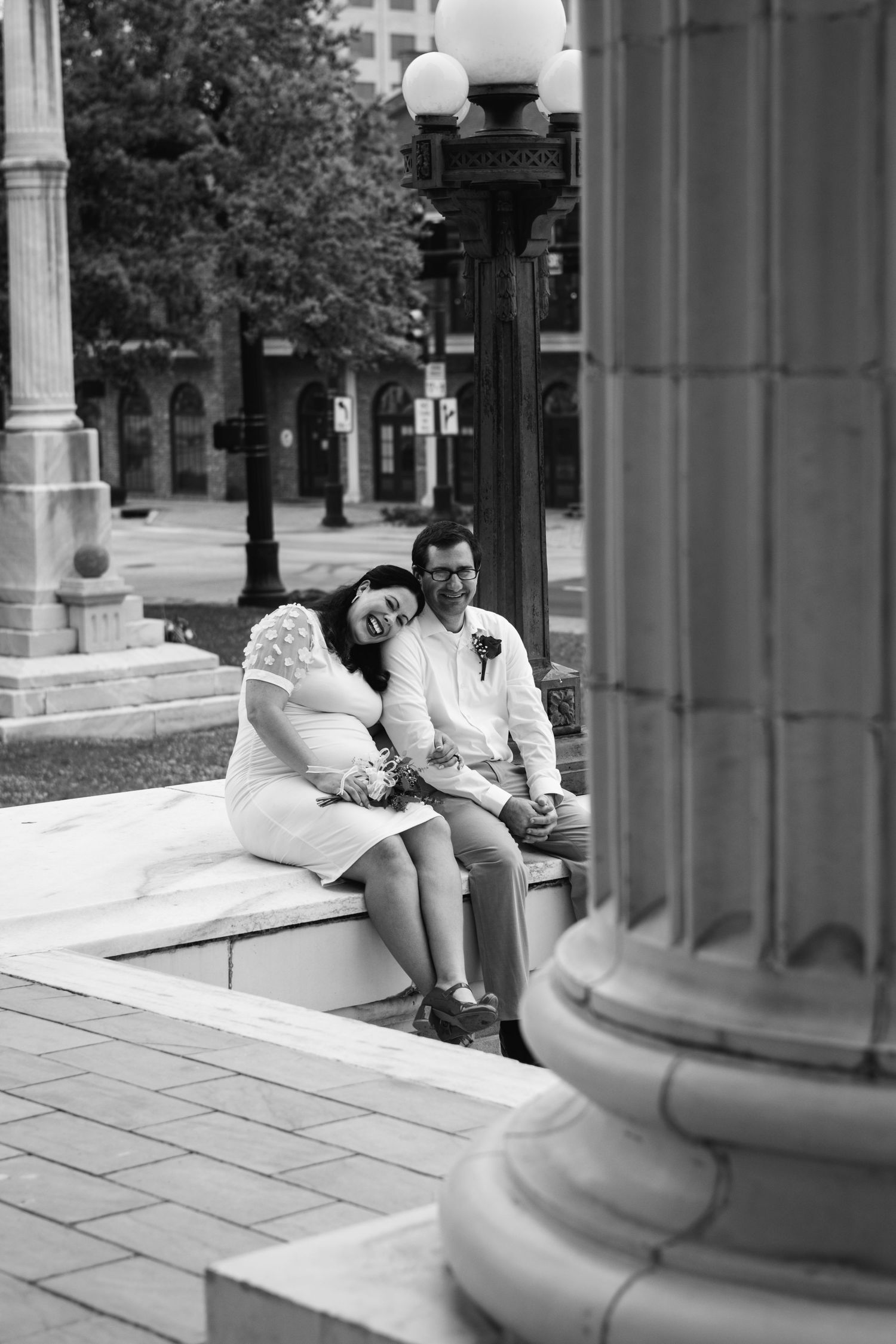 Black and white photo of a smiling couple sitting on steps near columns in an urban plaza, dressed in wedding attire.
