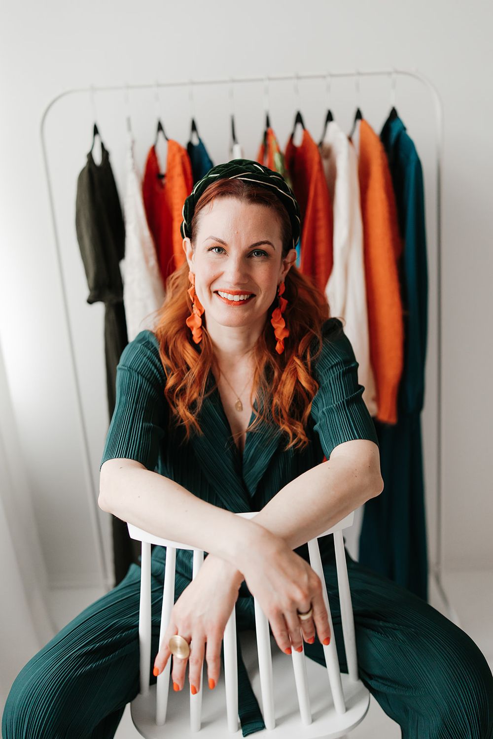 A woman in a green outfit sits on a white chair with clothing hanging on a rack behind her.
