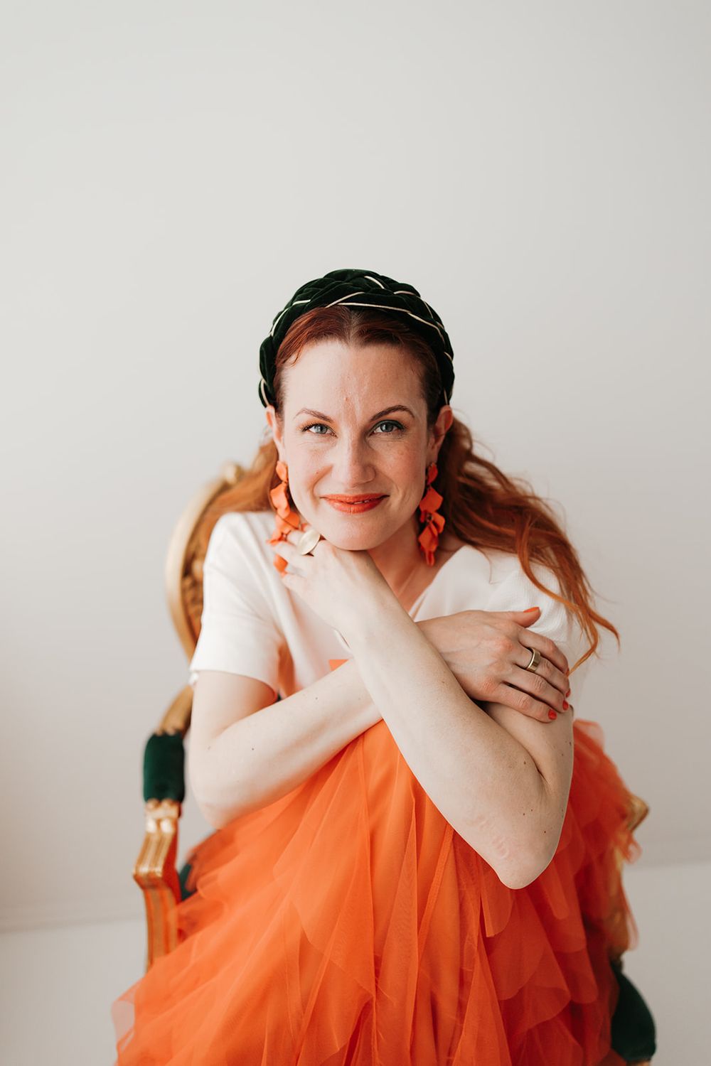 A person in a white top and flowing orange tulle skirt poses against a light background while wearing a green headpiece.