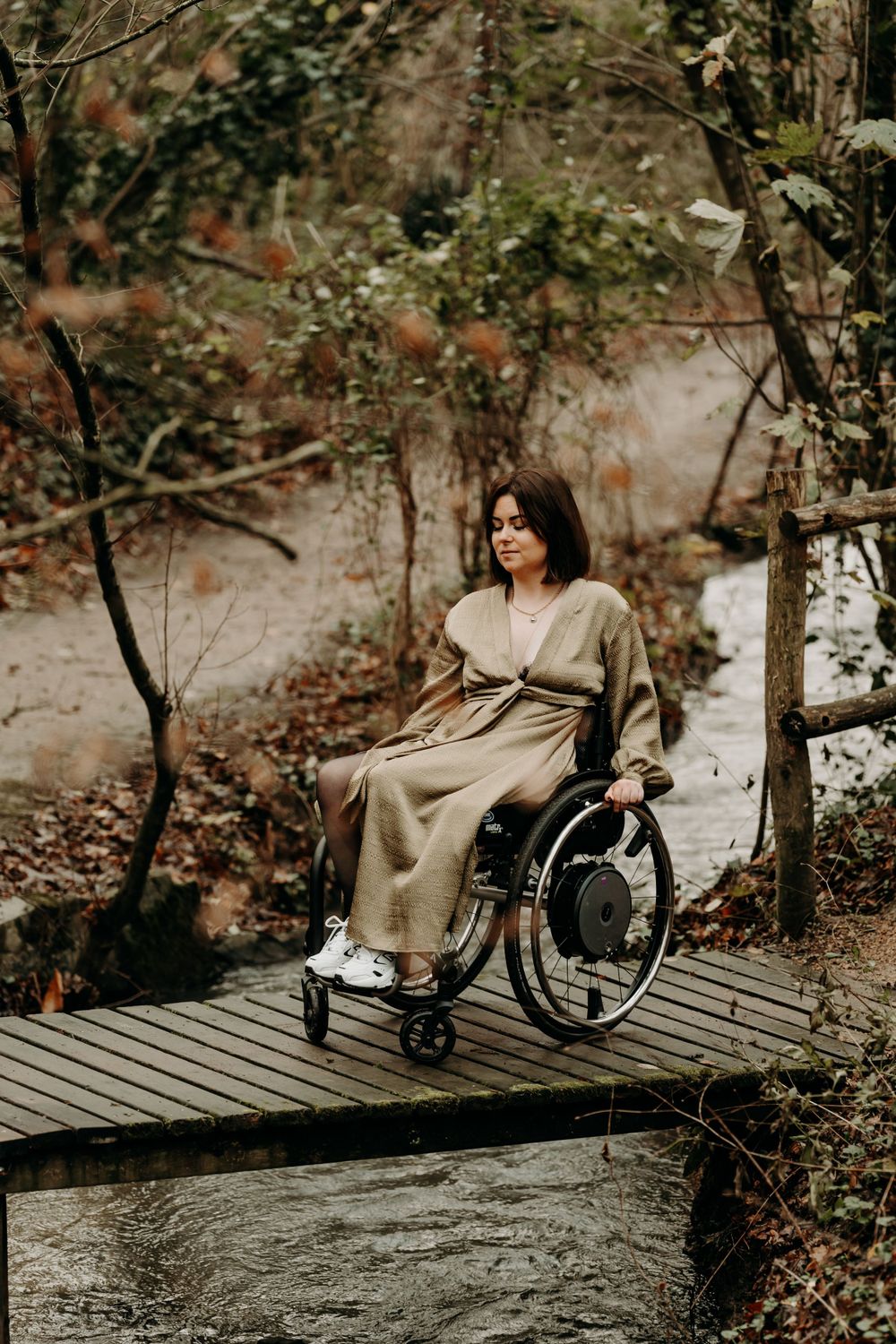 A person in a long dress sits in a wheelchair on a wooden pathway surrounded by autumn foliage in a forest setting.