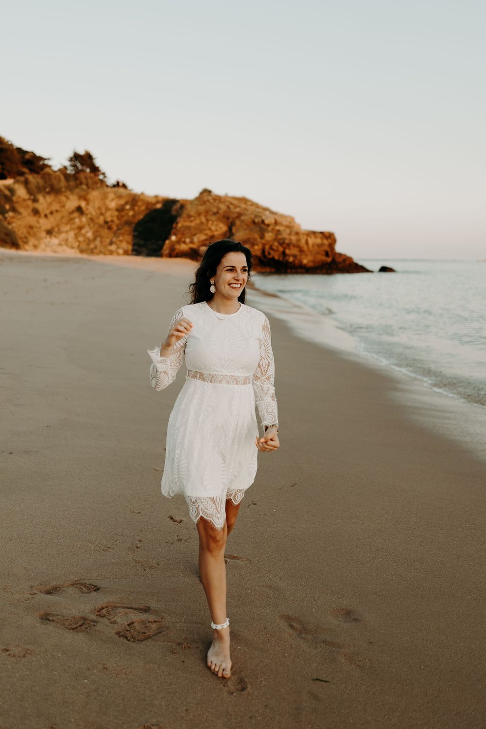 Someone wearing a white lace dress runs joyfully along a sandy beach at sunset with coastal cliffs in the background.