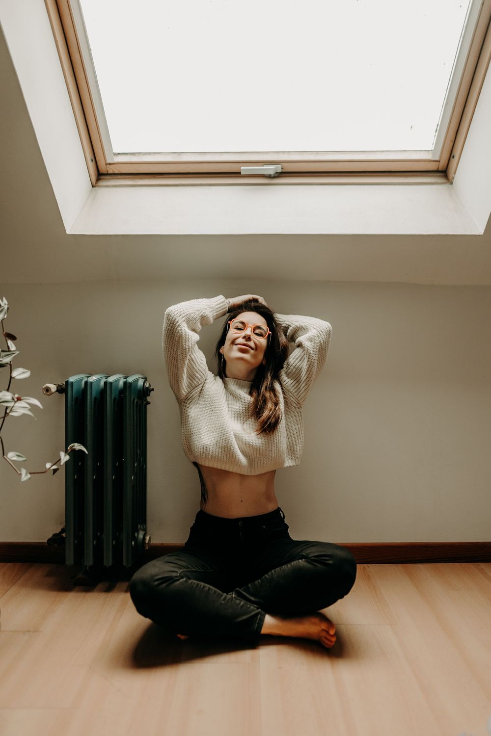 A person sits cross-legged on a wooden floor beneath a skylight, wearing a cream sweater and black pants.