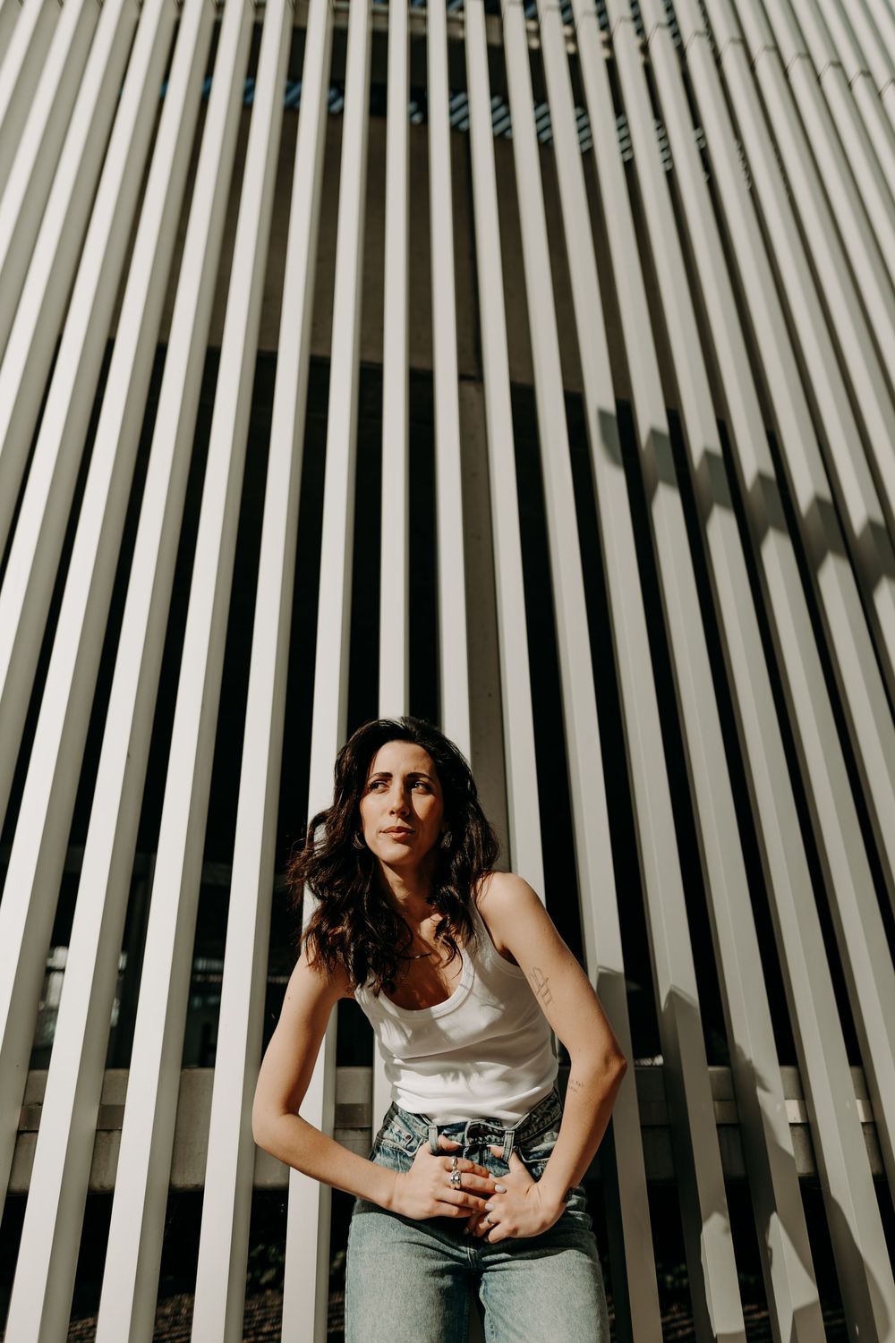 Woman in white tank top and jeans poses against striped architectural background with dramatic lighting and shadows.