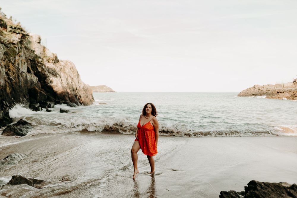 A person in a flowing red dress stands on a secluded beach between rocky cliffs at sunset.