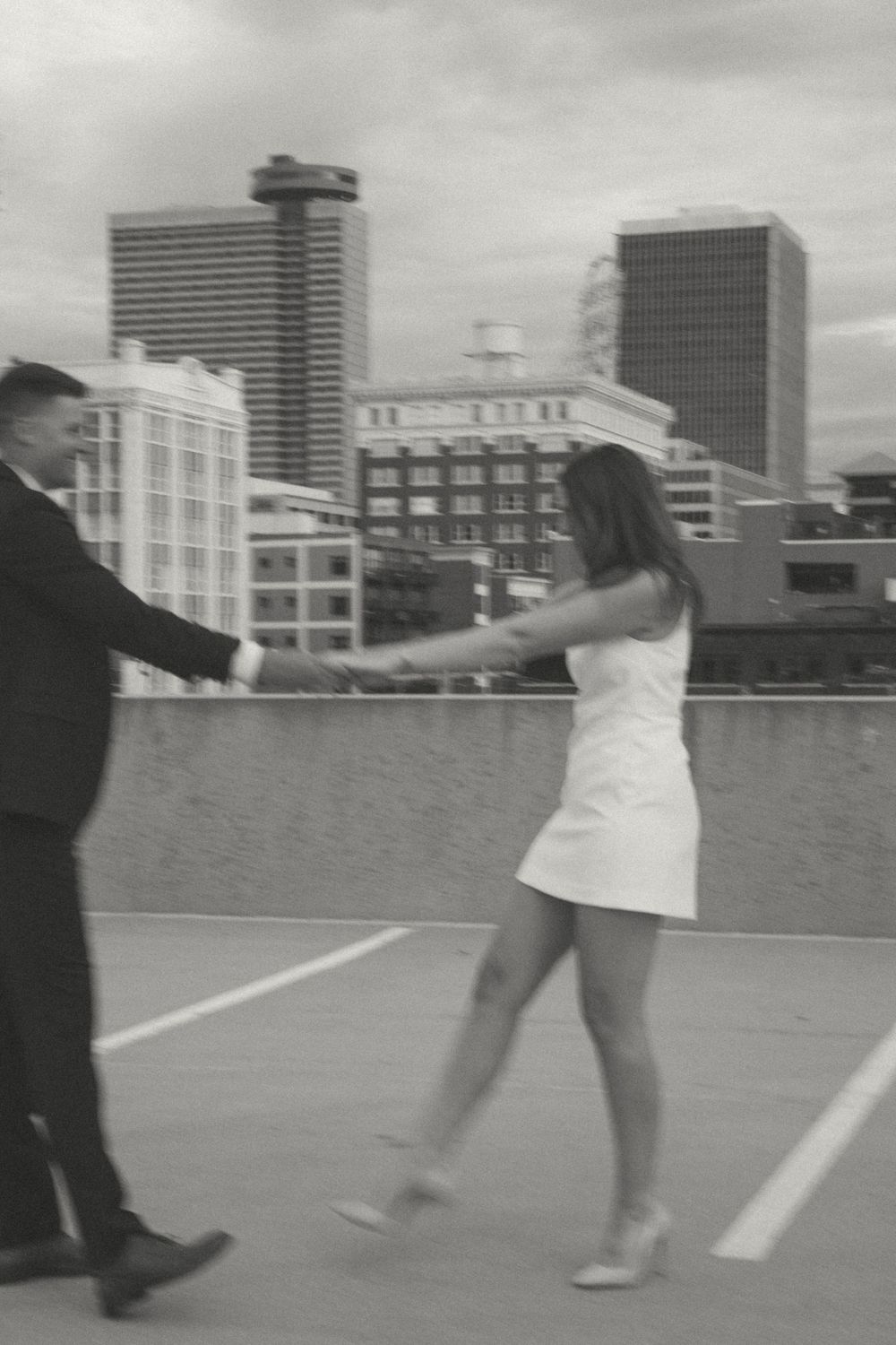 A couple dances together on a rooftop parking lot with city buildings in the background.