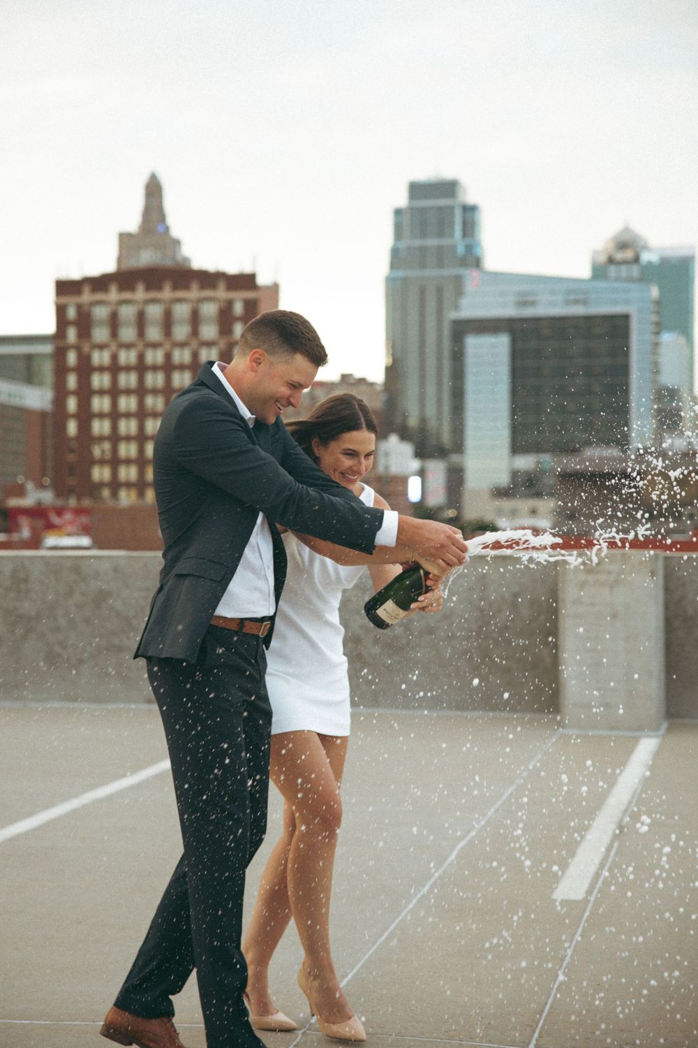 A couple celebrates by popping champagne on a city rooftop.