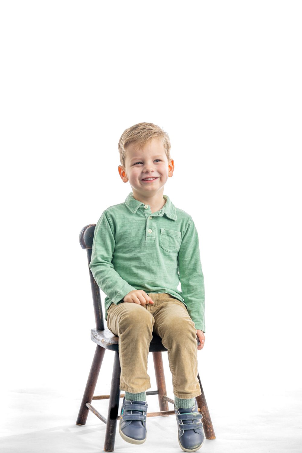 Child in green polo shirt and khaki pants sits on wooden chair against white background.