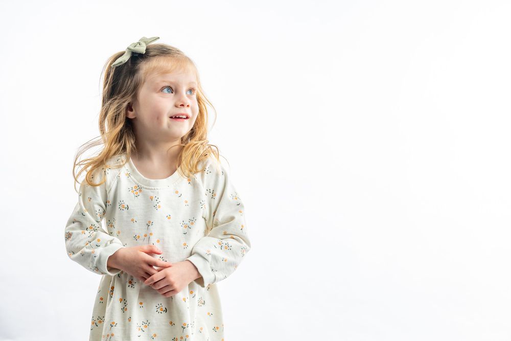 Young child wearing a white floral dress laughs while looking up on a white background.