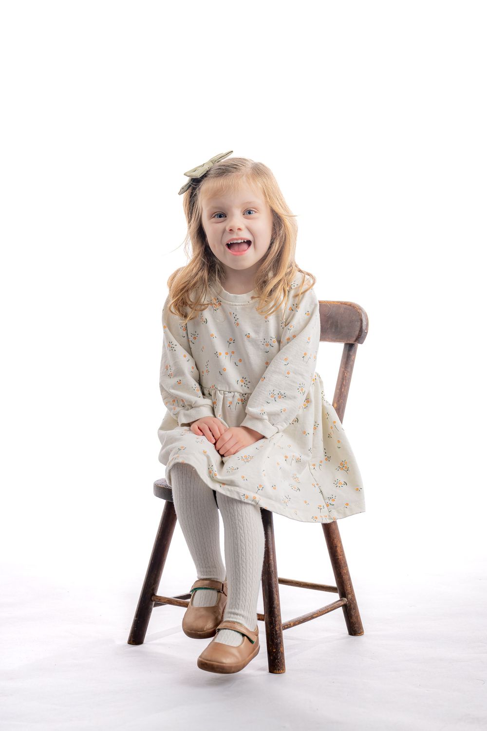 Small child wearing a cream-colored dress and tights sits on a wooden stool against white background.