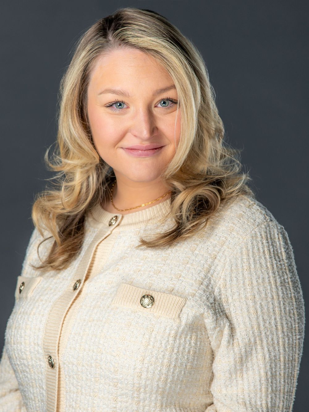 Professional headshot portrait of a person wearing a cream-colored textured jacket with gold buttons against a dark background.
