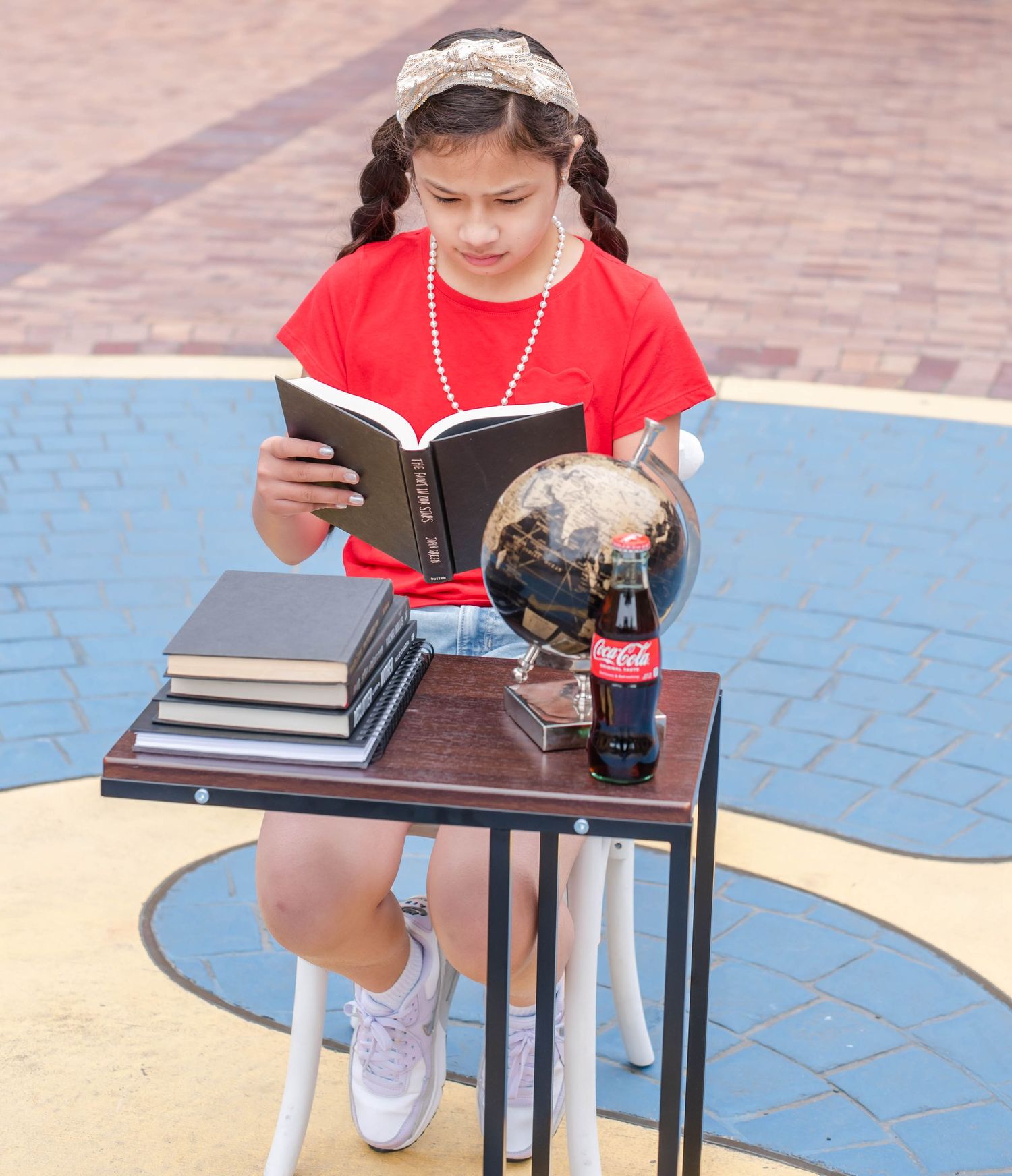Student reading from book while sitting at table outdoors in red t-shirt.