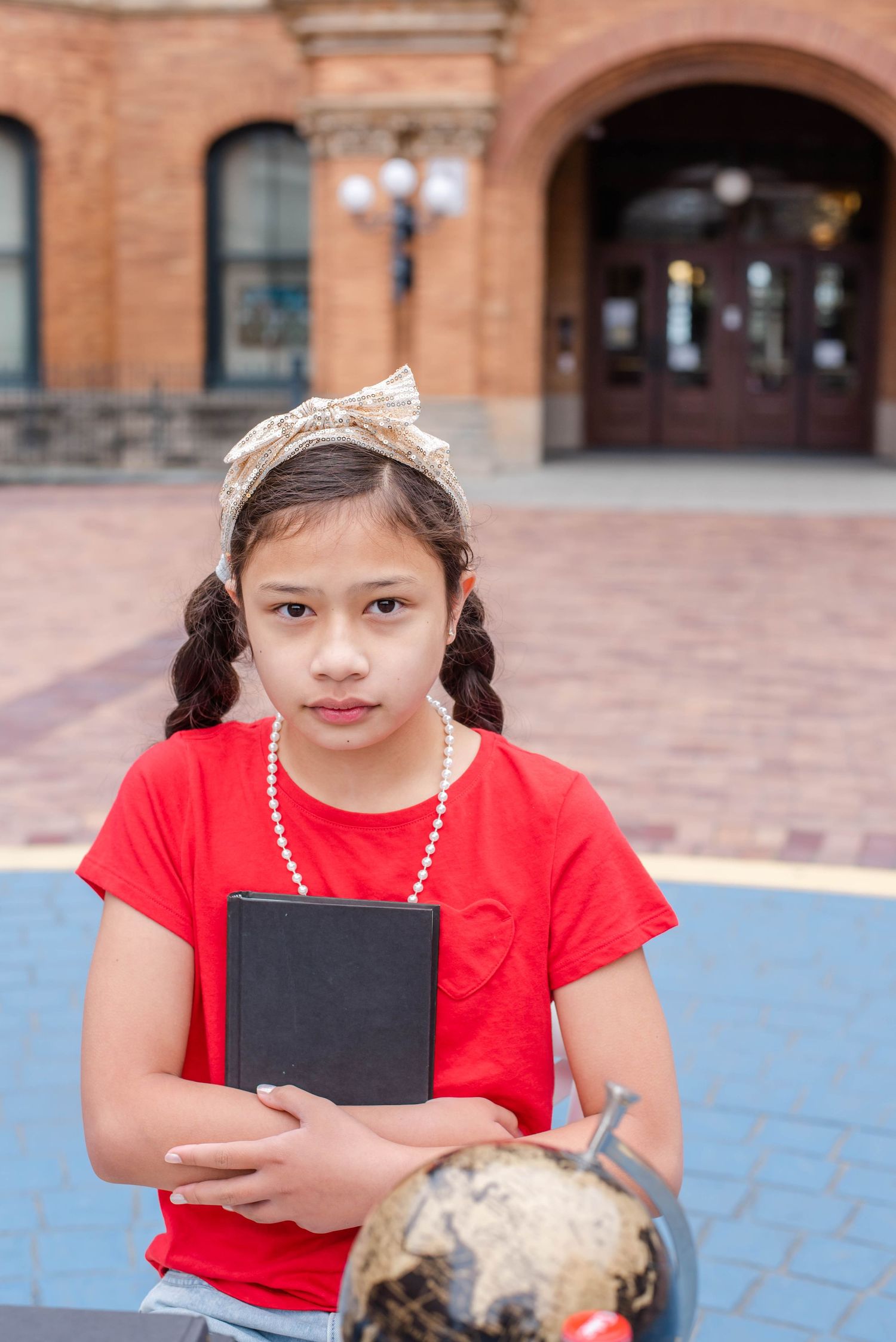Person in red t-shirt and pearl necklace reading outdoors near brick building.