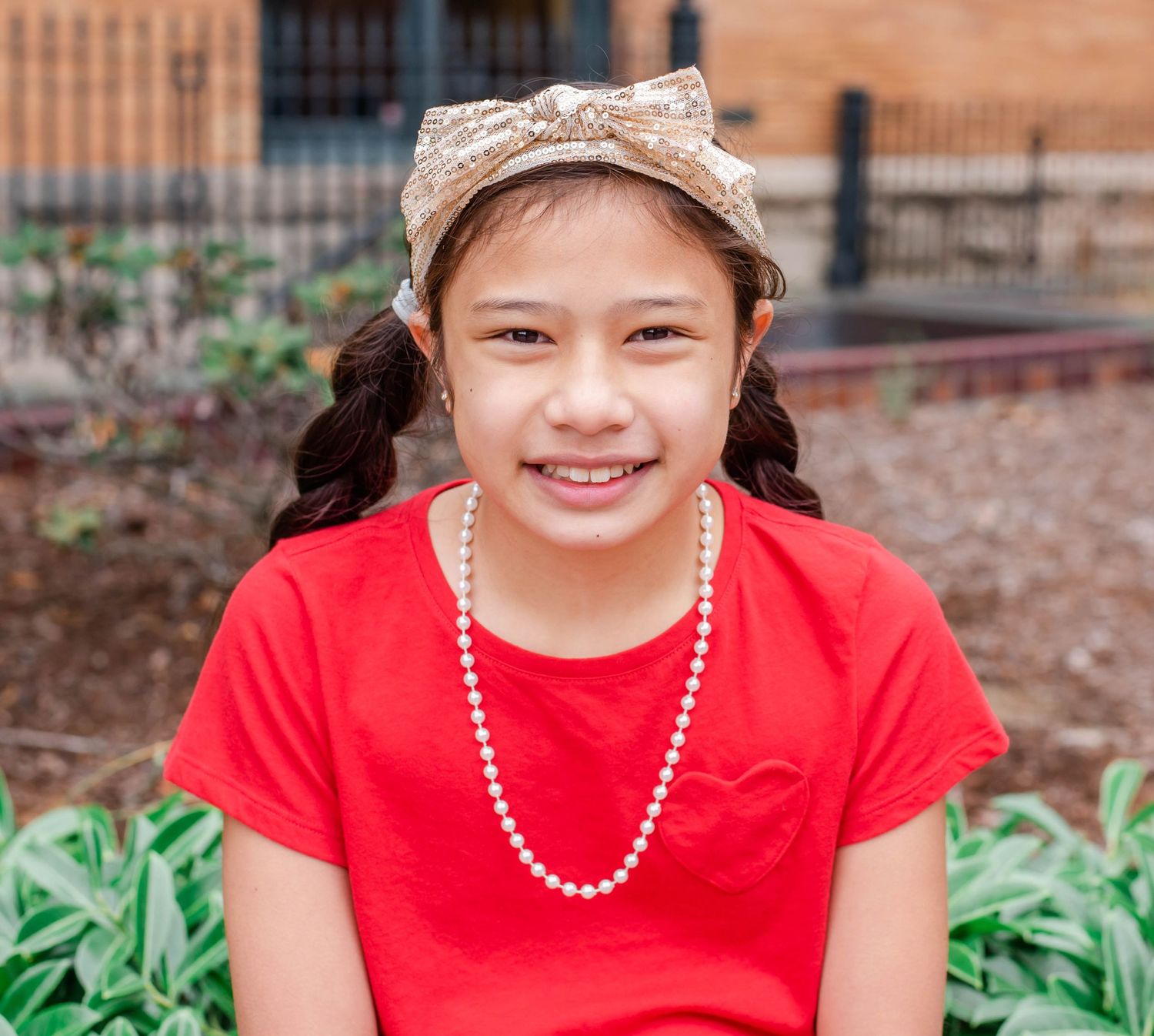 Portrait series against brick building wearing red shirt and pearl necklace.