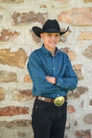 A young male wearing a cowboy hat and a blue shirt in front of a tan brick wall.