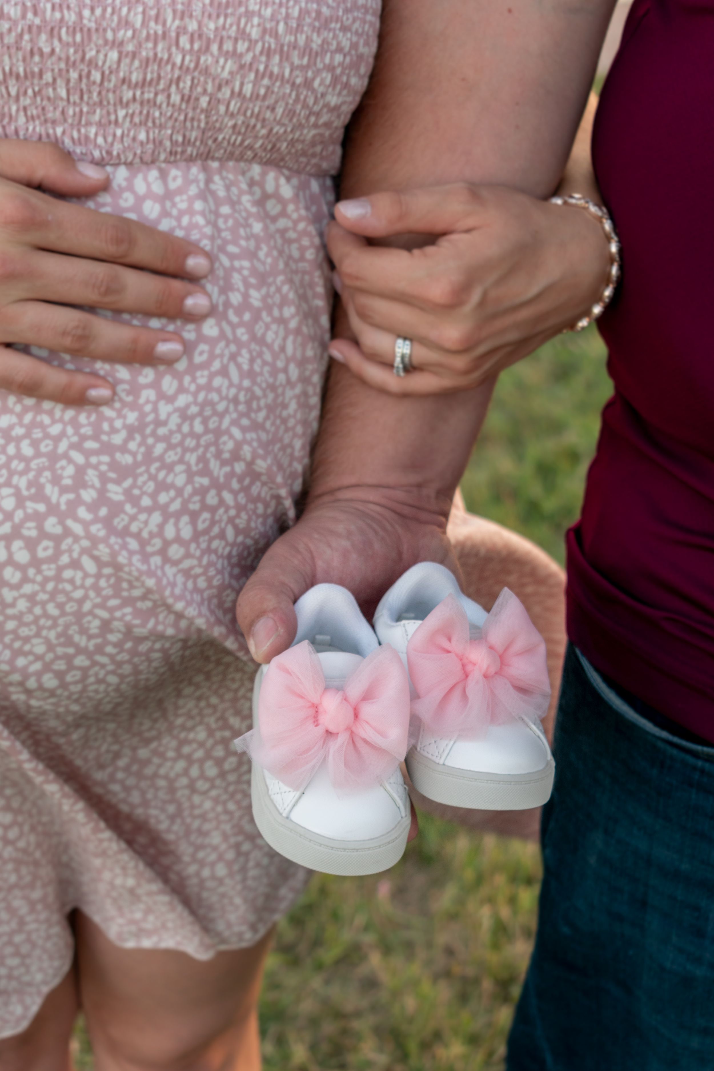A pregnant woman holding the arm of a man. The man is holding a pair of white baby shoes with pink bows.