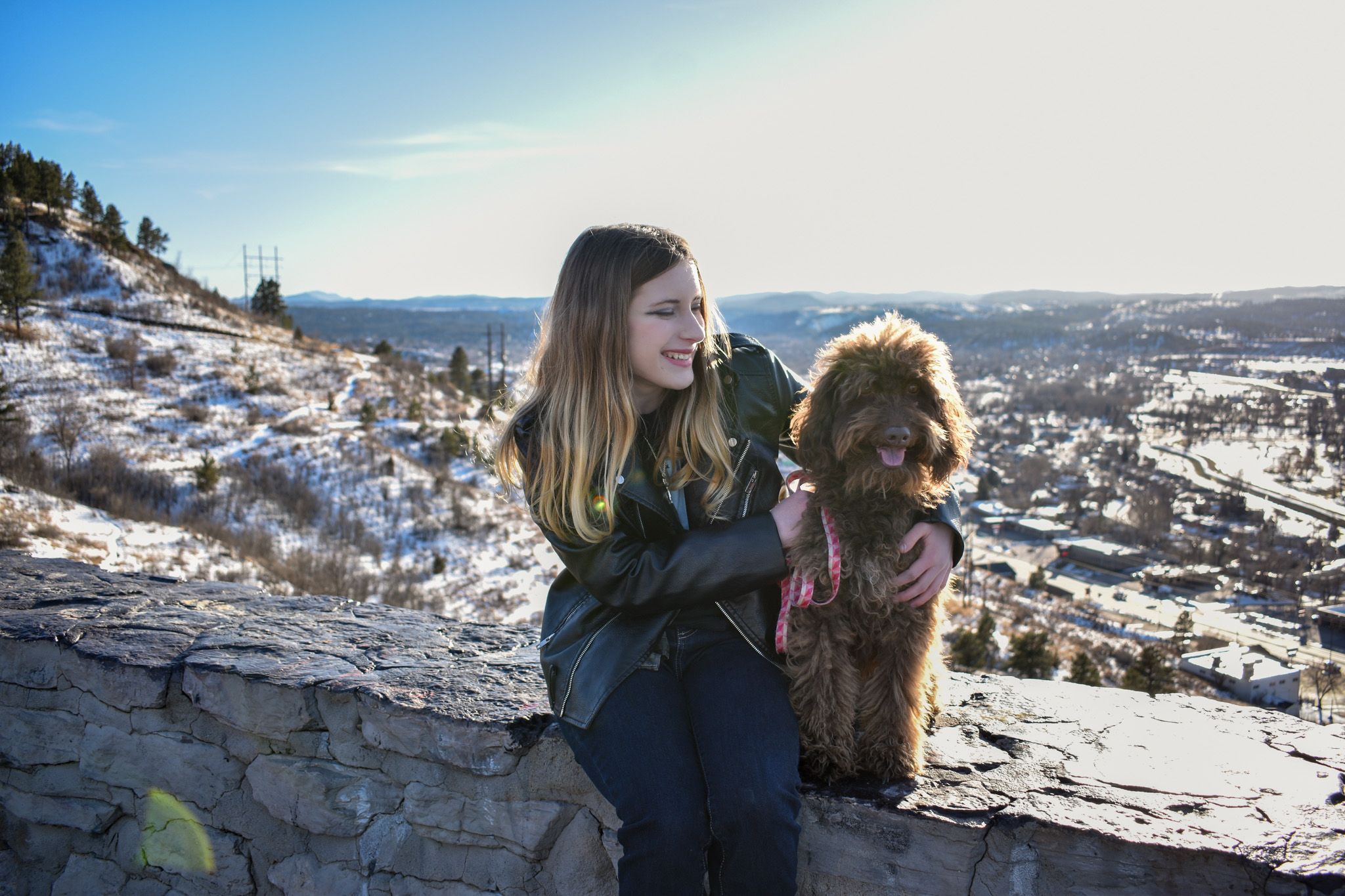 A girl and her dog sitting on a rock wall with a mountainous background