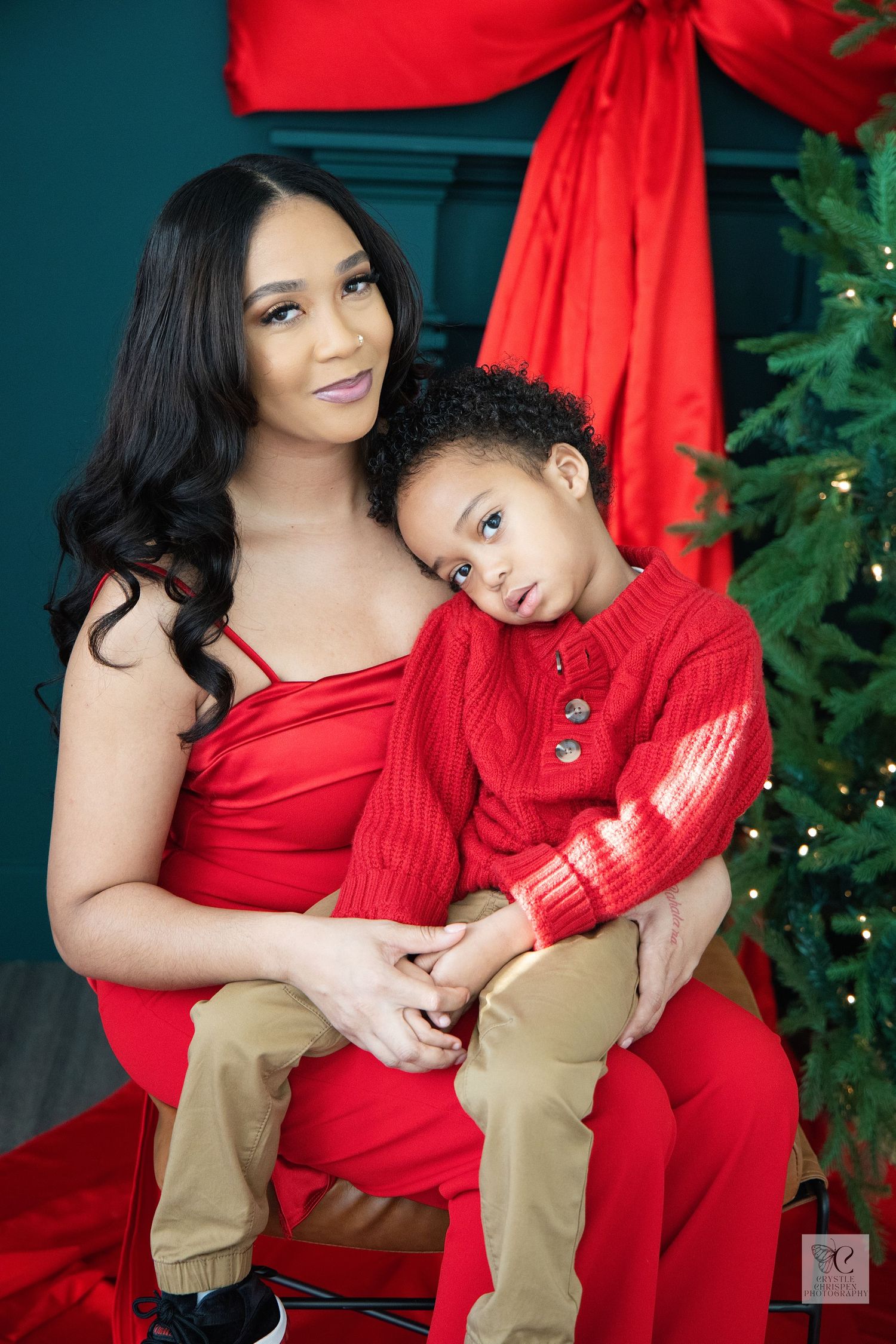 A mother and child wearing matching red outfits pose together for holiday photos in front of a Christmas tree.