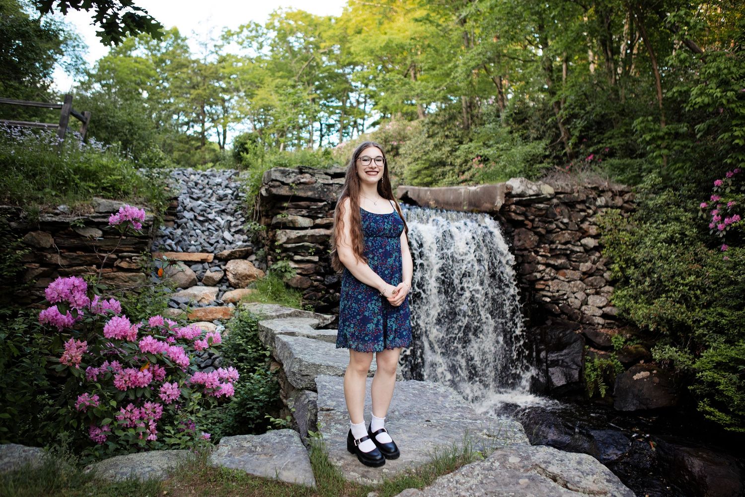 High school senior girl posing in front of waterfall, stone wall and flowers.