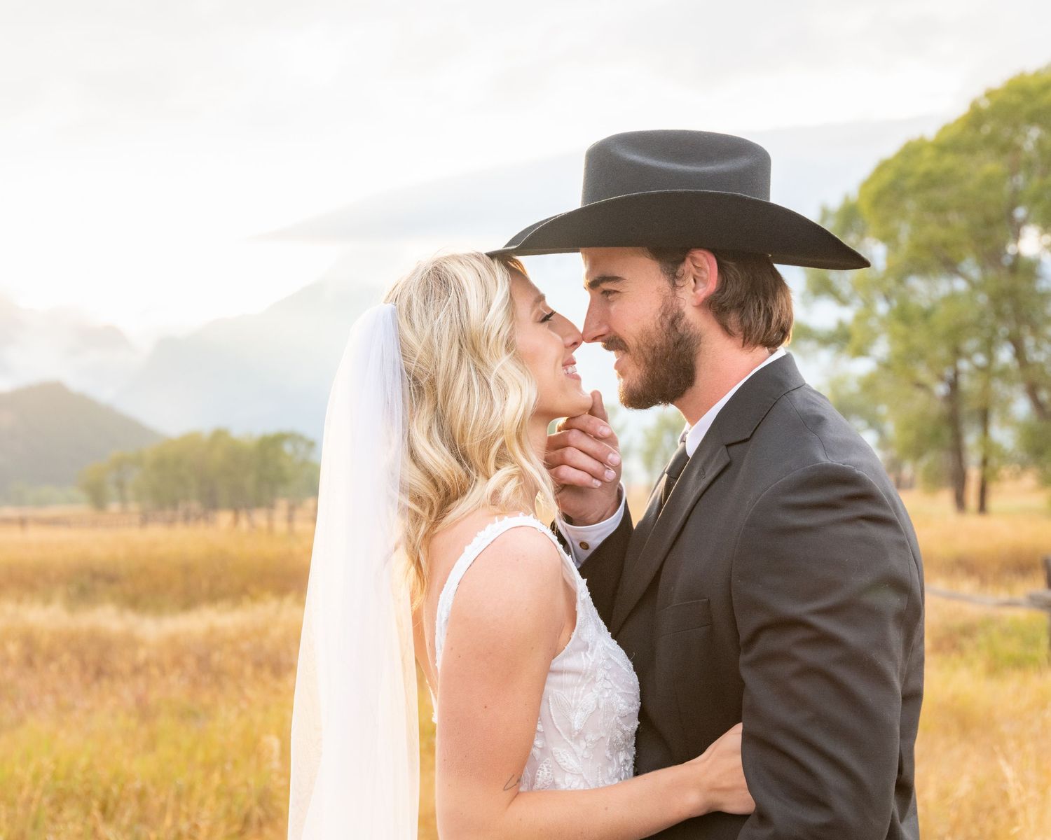 A western-styled couple shares a romantic moment in a golden field with mountains behind them.