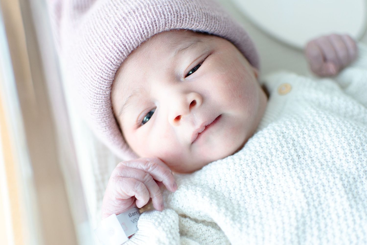 Smiling newborn baby wearing a soft pink knit hat and white swaddle.