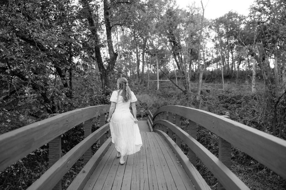 Black and white photo of a person in white dress walking across a wooden bridge surrounded by trees.