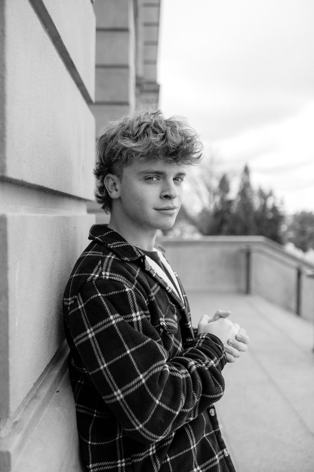 Black and white portrait of someone in a plaid flannel jacket leaning against a wall outdoors.