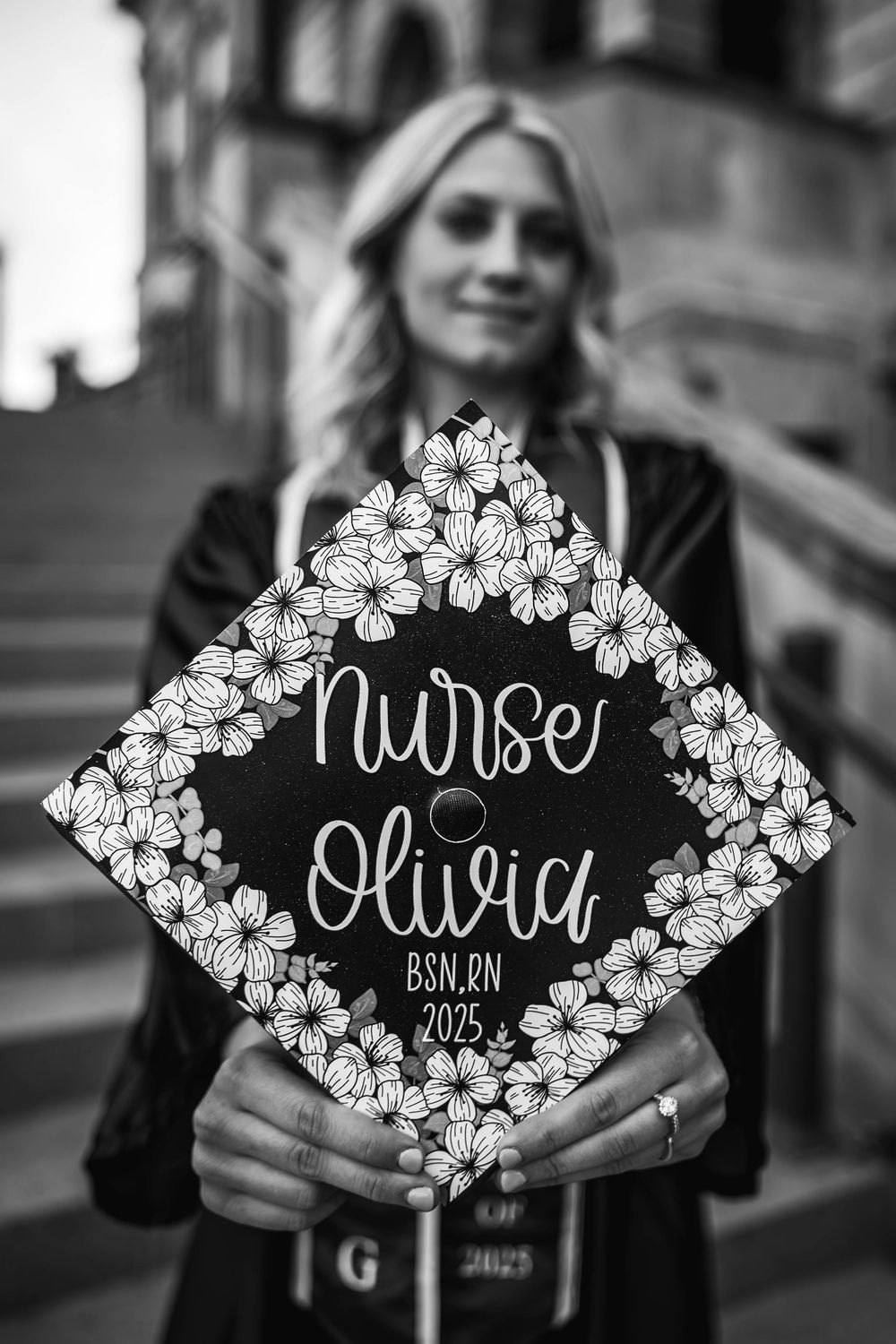 A decorated graduation cap with 'Nurse Olivia' written in white script surrounded by floral designs on black background.