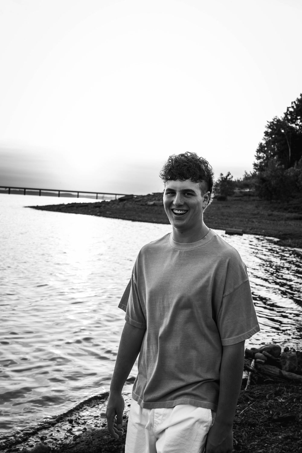 Black and white portrait of person by waterfront wearing oversized t-shirt with dock in background.