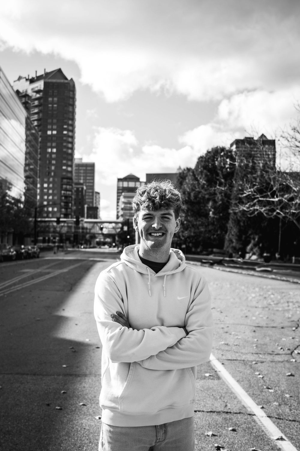 High school senior in white hoodie stands on urban path with tall buildings and cloudy sky in black and white photo.