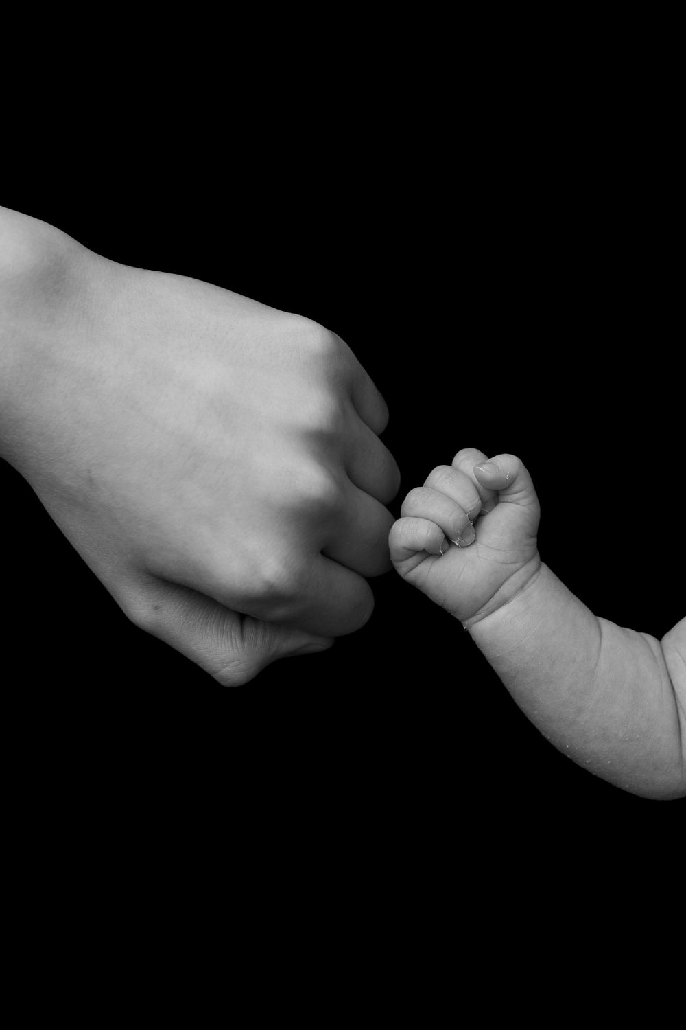 Black and white image of adult and baby hands fist-bumping against dark background.