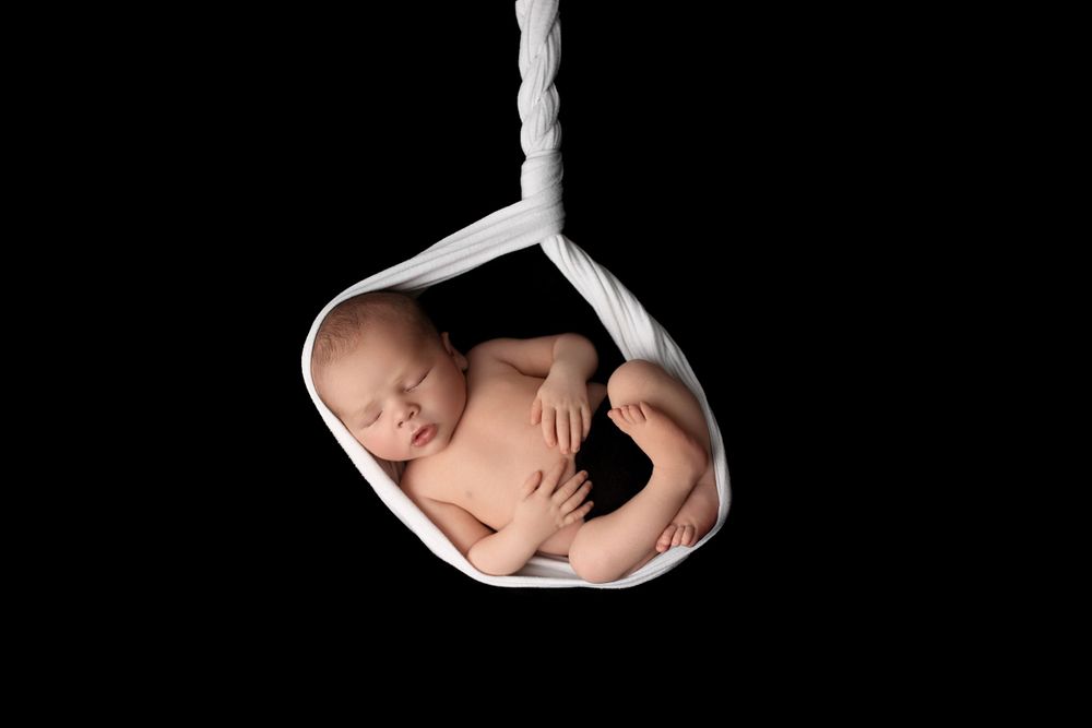 Sleeping newborn baby curled up in white fabric sling against black background.